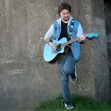 music performer leaning against a wall holding a guitar